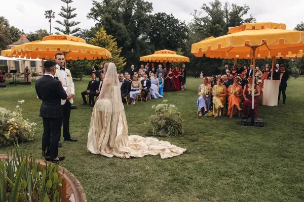 Wedding ceremony with couple exchanging vows as bride’s cathedral veil and long train trail down the aisle on a garden lawn under umbrellas