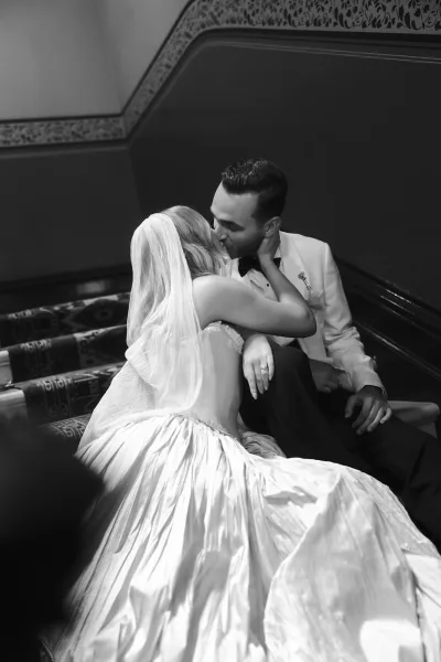 Wedding kiss portrait in black and white of bride and groom kissing on a staircase, her long veil draped over his tuxedo in a hallway interior