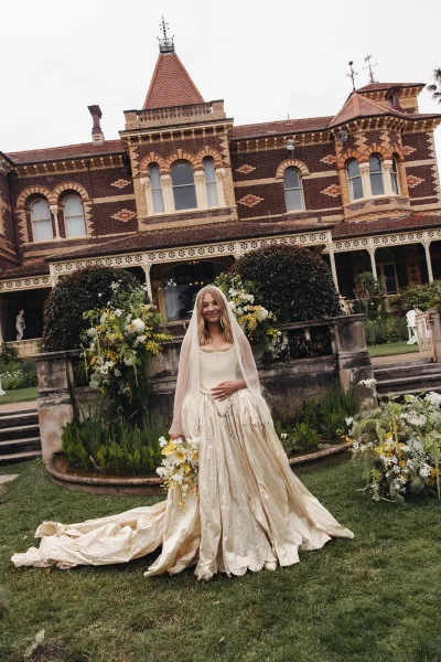 Bridal portrait of a bride holding bouquet, cathedral veil trailing from her champagne satin gown beside a stone fountain at a brick estate garden