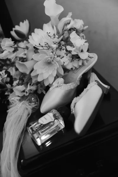 Bridal shoes in a wedding shoes flatlay with bouquet accent, perfume bottle, jewelry, and veil arranged on a dark tabletop