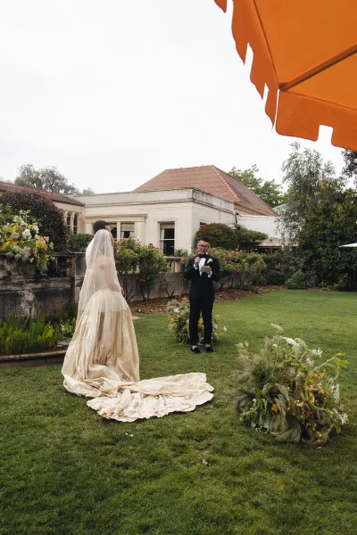 Wedding vows as groom reads from a card into a microphone while bride in cathedral veil and long train faces him amid garden florals