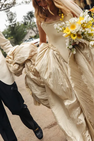 Bride and groom walking as bride holds bouquet of white and yellow calla lilies, groom lifting her dress train on a tree-lined walkway