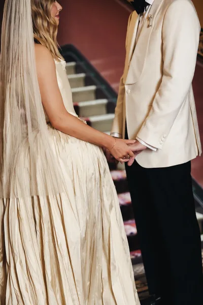 Wedding couple portrait of bride and groom holding hands on an indoor staircase, her long veil and strapless gown beside his ivory tuxedo jacket