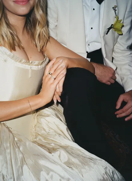 Couple portrait of bride and groom close up, her off-shoulder satin dress and rings as she holds his arm in a white tux against a dark backdrop