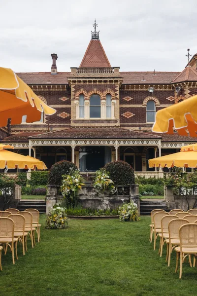Ceremony setup with outdoor ceremony seating of cane-back chairs lining a lawn aisle, yellow and white florals by a brick facade veranda