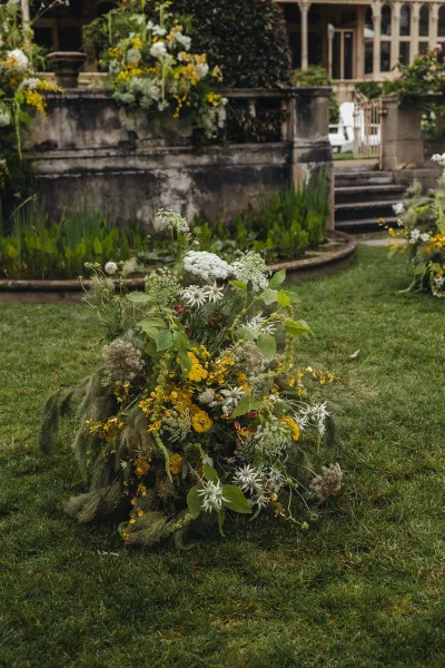 Wedding floral installation with grounded wedding flowers in yellow and white wildflowers and greenery beside a stone fountain on a lawn