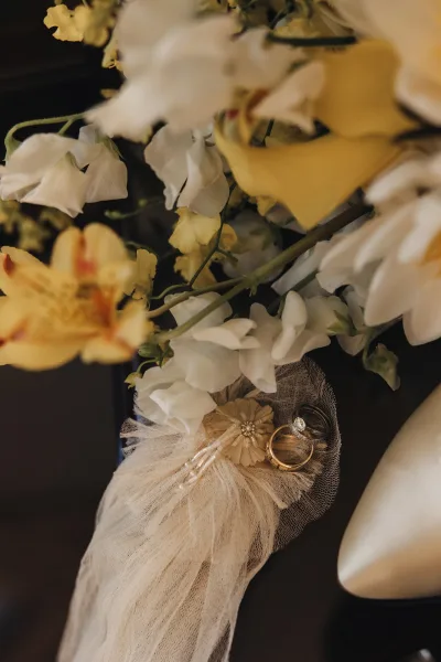 Wedding rings with an engagement ring close up, resting on tulle ribbon beside a floral bouquet with pearl detail on a dark surface