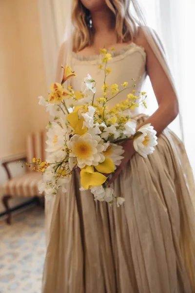 Bridal bouquet of yellow and white bouquet flowers tied with ribbon, held by bride in a spaghetti strap dress by window light indoors