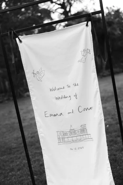 Wedding welcome sign on a metal stand, a fabric banner with grommets and ribbon ties, featuring line art on a lawn with trees