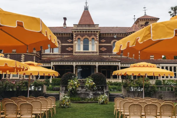 Outdoor ceremony setup with wedding ceremony chairs in rattan beneath yellow patio umbrellas, floral steps leading to a brick veranda backdrop