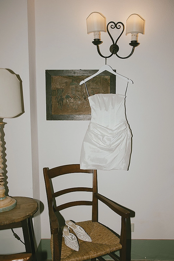 Wedding dress hanging on a hanger beside bridal shoes on a wooden chair in a softly lit room with a white wall and sconce