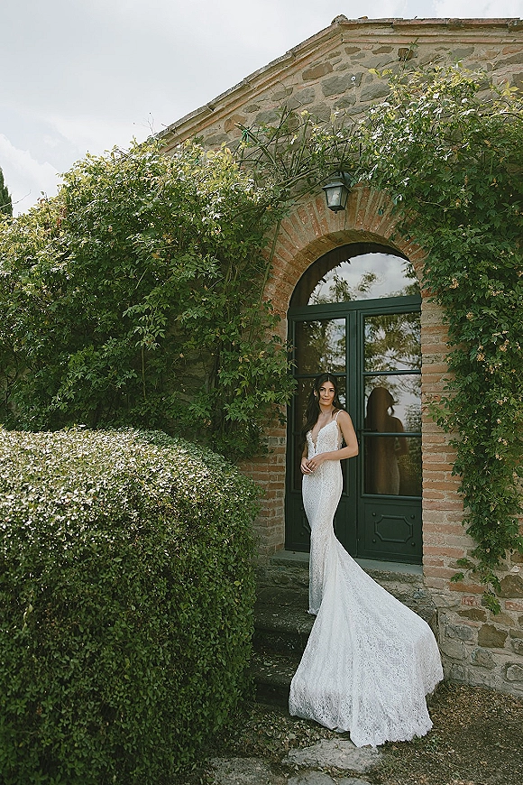 Bridal portrait of a bride in a lace wedding dress with long train, standing in an ivy-framed stone doorway by a green door