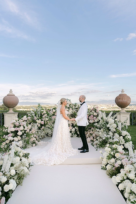 Wedding ceremony moment as bride and groom holding hands on an outdoor terrace platform, with rose-lined aisle, stone balustrade, and mountain view