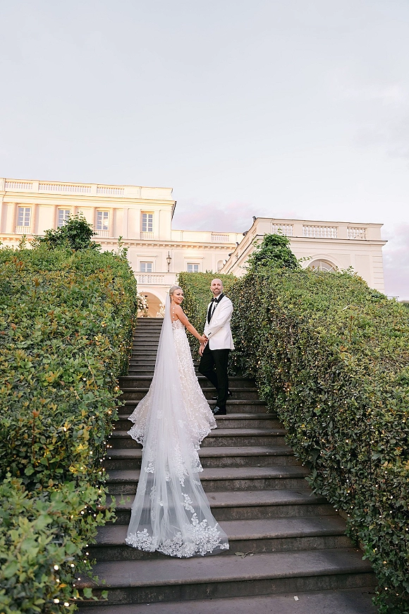 Couple portrait of bride and groom holding hands on a stone staircase, her long veil trailing under string lights by a grand white estate at dusk