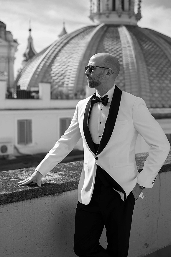 Groom portrait in a white tuxedo, wearing sunglasses and a black bow tie, leaning on a rooftop stone ledge with skyline view