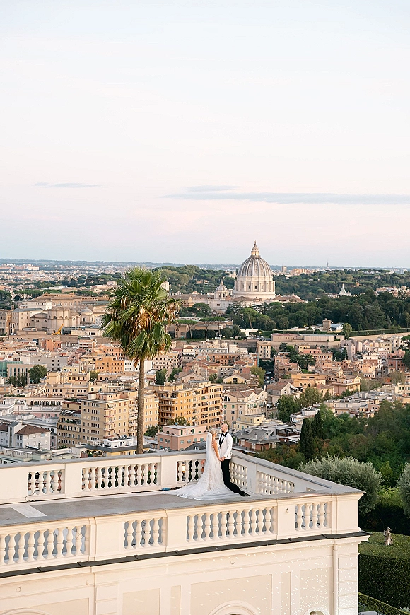 Couple portrait of bride in lace gown with long veil and groom in tuxedo holding hands on a rooftop terrace with city skyline and cathedral dome
