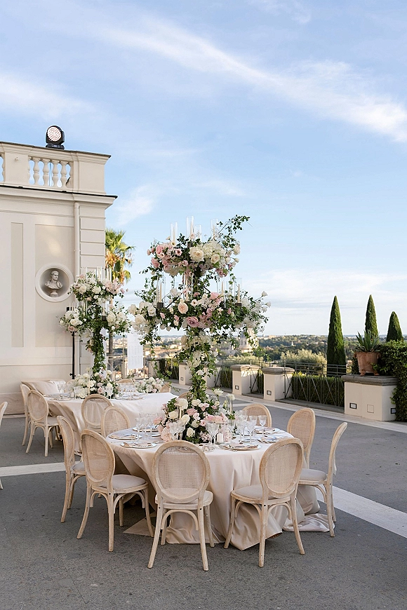Reception tablescape with an outdoor wedding reception table set on an outdoor terrace, ivory linens, roses, candles, and cane chairs by a white facade