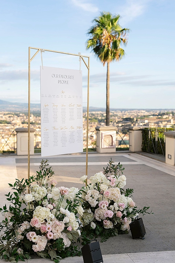 Wedding seating chart on a gold frame with ribbons and white and blush florals, set on an outdoor terrace with palm tree and skyline backdrop