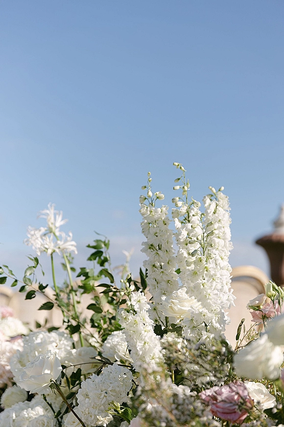 Wedding florals with white wedding flowers, delphinium, roses, and hydrangea with greenery, set outdoors under blue sky by a blurred fountain