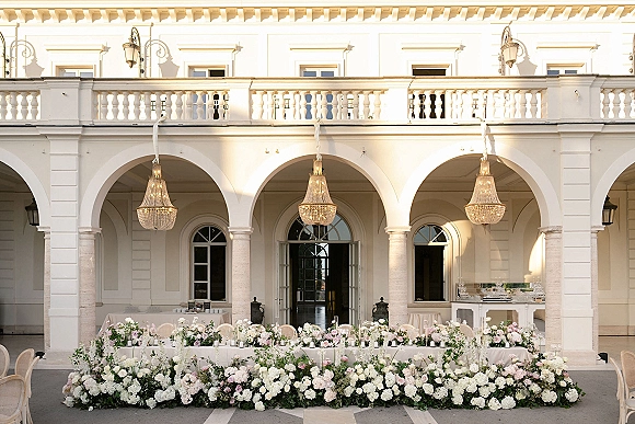 Wedding head table with head table floral garland, white taper candles and chandeliers beneath an arched courtyard colonnade backdrop