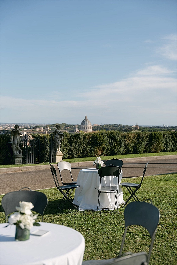Outdoor reception tables with round wedding table setup, white linens, black chairs, and bud vase centerpieces on a lawn with skyline backdrop
