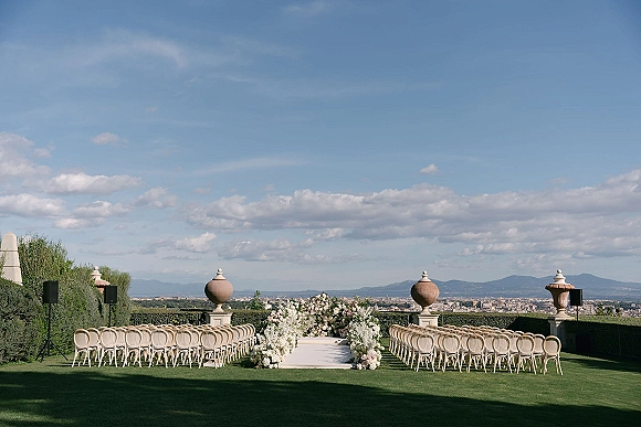 Ceremony setup with a white aisle runner and blush florals lining the aisle, wooden chairs on a lawn with mountain and city views