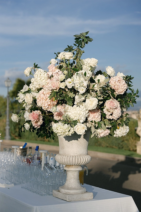Wedding floral arrangement in a stone urn with white and blush roses and hydrangeas on a bar table, with glassware and blue sky behind