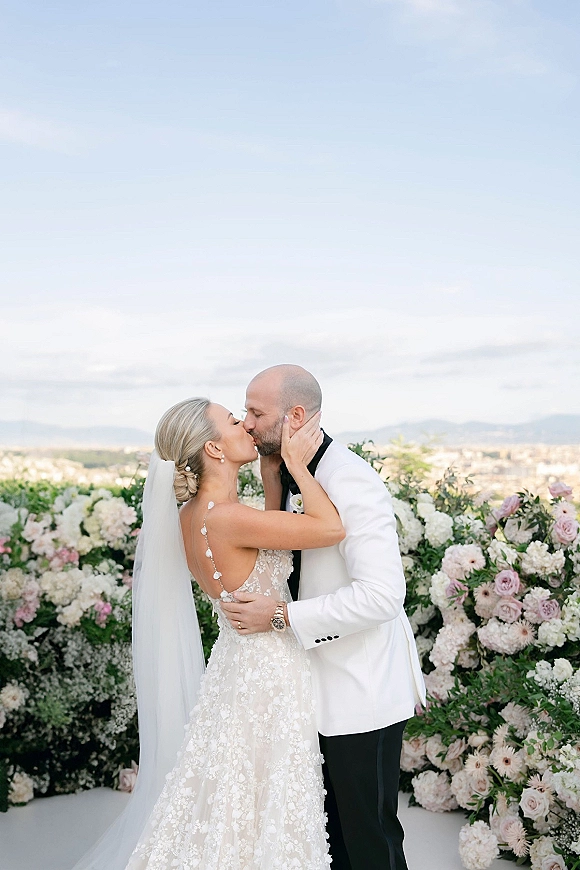 Wedding kiss portrait of bride and groom kissing as she holds his face, veil flowing, with floral wall backdrop on a terrace with city and mountains view