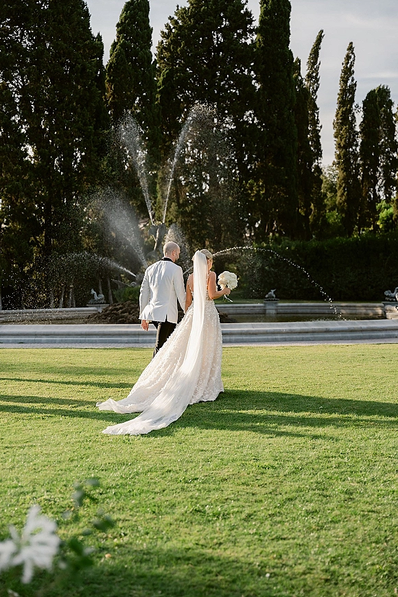 Couple portrait of bride and groom walking away hand in hand, her long veil and dress train trailing on grass by a fountain in a garden
