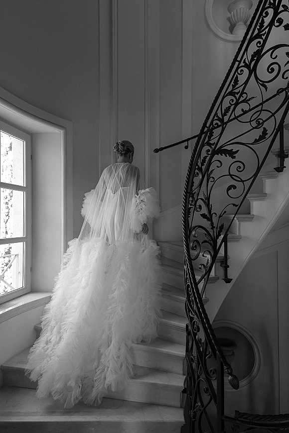Bridal portrait of a bride in a feathered wedding dress and tulle veil, posed on a grand staircase with wrought iron railing and window light