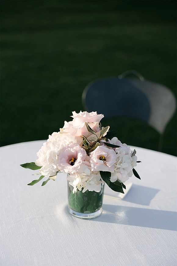 Wedding centerpiece with pink and white flowers and greenery in a glass cylinder vase on a white tablecloth, set on a green lawn near a chair