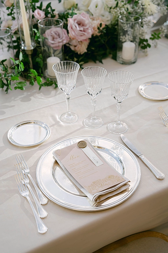 Reception place setting with a silver charger plate, wax-sealed menu and place card, crystal stemware, and candlelit florals on a white tablecloth