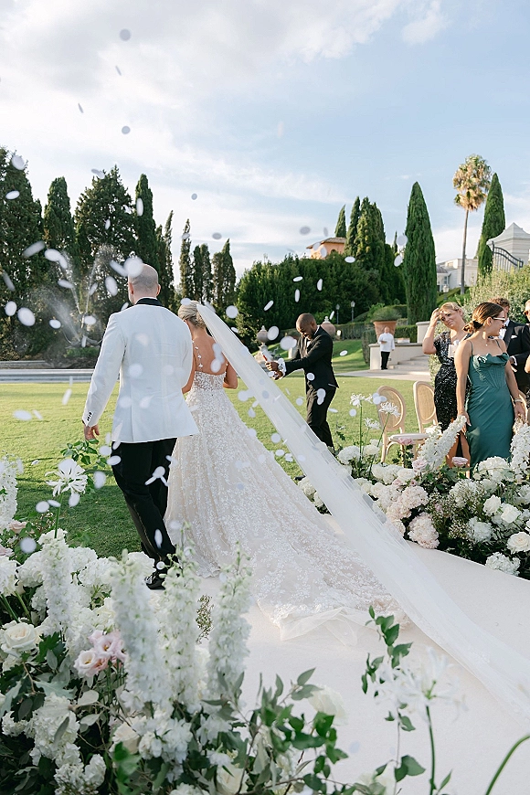 Wedding recessional with flower petal toss as newlyweds walk away, bride’s long veil trailing behind, groom in white jacket on garden lawn aisle