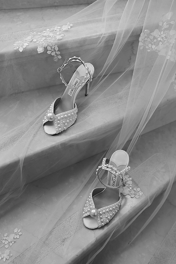 Bridal shoes with pearl wedding shoes details, ankle-strap open-toe heels beside a tulle veil with lace appliqués on stone stairs