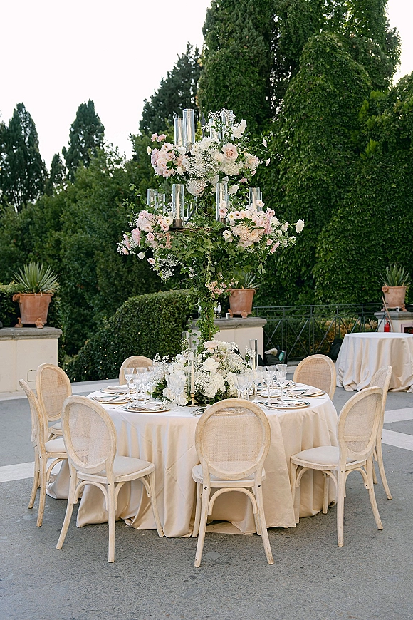 Reception tablescape with round wedding table decor featuring tall blush and white roses, hydrangea, candles, and gold flatware on a terrace