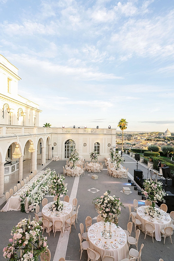 Outdoor wedding reception set with round tables in blush linens, floral centerpieces and candlelight beneath chandeliers on a terrace courtyard