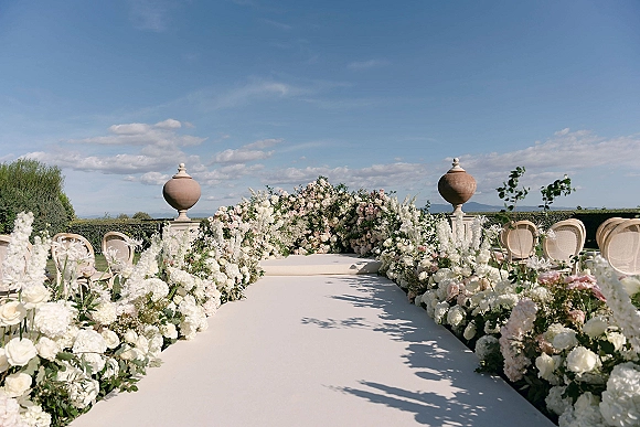 Ceremony aisle decor with a white aisle runner, floral ground arrangements of white and blush roses by wicker chairs on a garden terrace