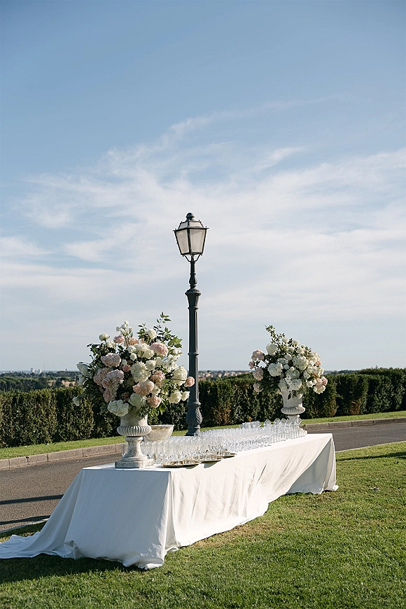 Wedding cocktail table with outdoor champagne coupe display, rows of glasses and large urn florals on white linen on a sunny lawn