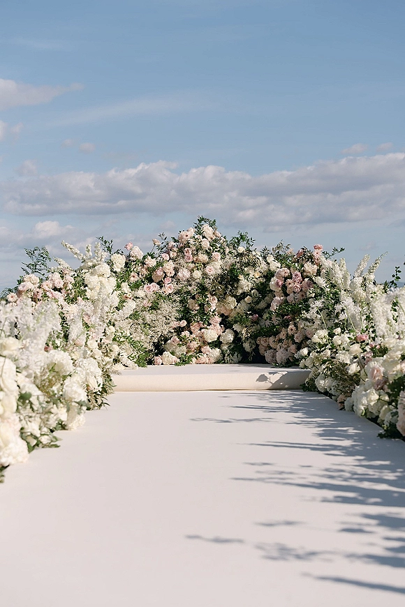 Wedding ceremony backdrop with a floral ceremony arch of blush and white roses, greenery, and ground florals beneath a blue sky