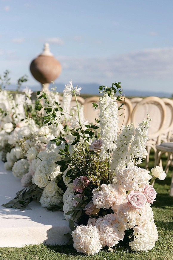 Ceremony aisle flowers line a fabric runner, with white florals and blush roses beside chairs on a lawn with distant hills.