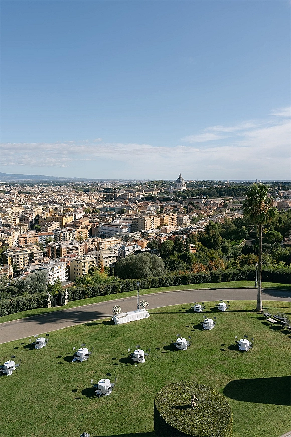 Outdoor reception setup with round tables in white tablecloths and floral centerpieces on a lawn, with statues and city skyline beyond