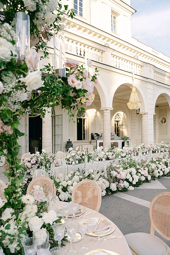 Reception tablescape at an outdoor wedding reception with blush roses, hydrangeas, taper candles, and rattan chairs under villa arches