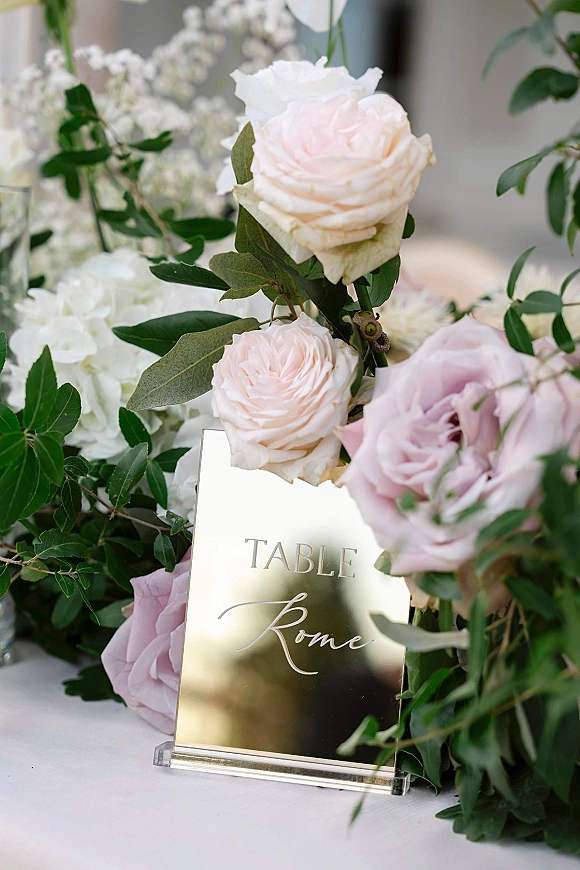 Wedding table sign with a mirror table number sign in gold, set in an acrylic holder with blush and white roses, hydrangeas, greenery on white linen