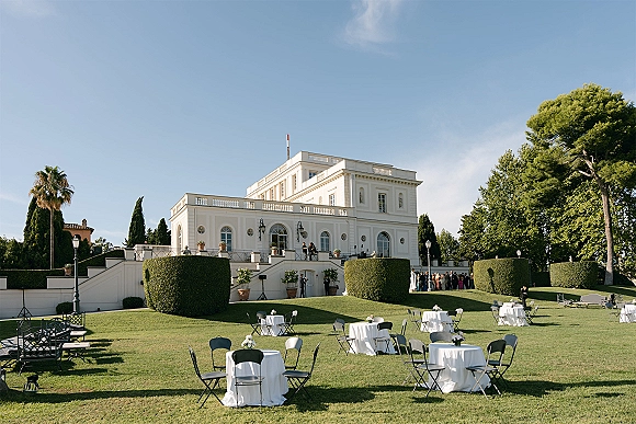 Outdoor wedding reception with garden reception layout of round tables in white linens and bistro chairs on a manicured villa lawn under blue sky
