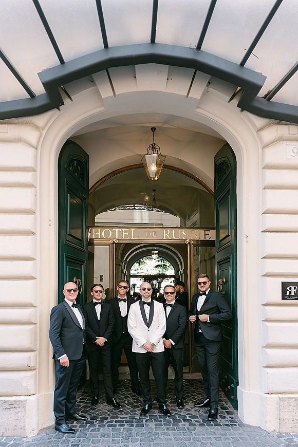 Groomsmen portrait of groom in a white dinner jacket with black tie groomsmen in tuxedos and sunglasses at a hotel entrance doorway