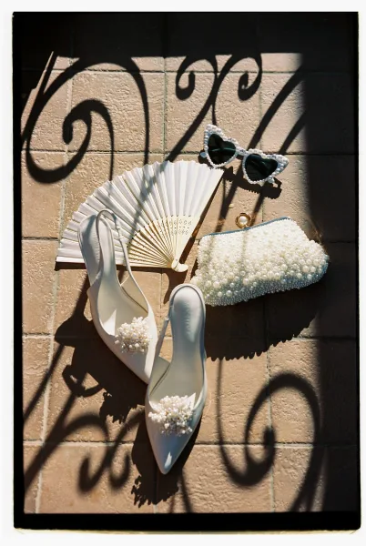 Bridal accessories flatlay featuring wedding shoes flatlay with pearl slingback heels, beaded clutch, heart sunglasses, fan, and gold ring on sunlit tiles with wrought iron shadows