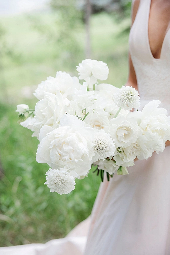 Bridal bouquet of white wedding bouquet blooms with peonies, ranunculus and scabiosa, held against a bridal gown in a green field