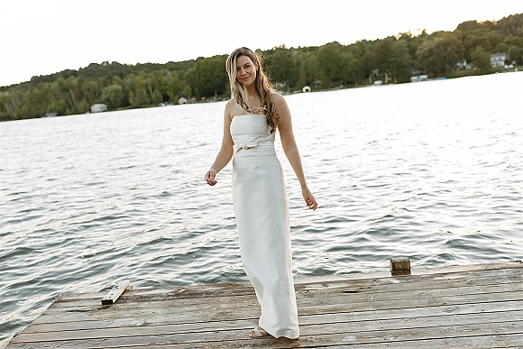 Bridal portrait of a bride in a strapless wedding dress with a sash, standing on a wooden dock by a calm lake and forested shore