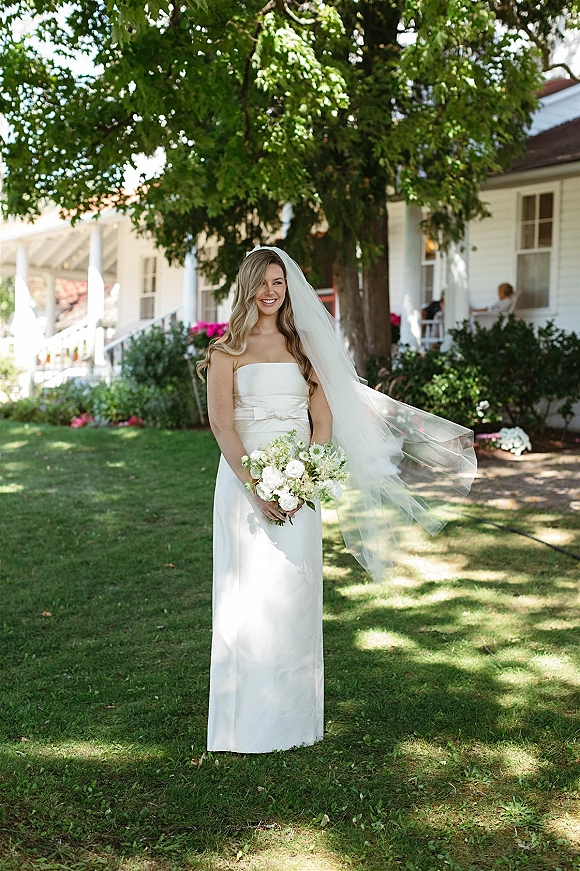 Bridal portrait of a bride holding bouquet in a strapless wedding dress, long veil blowing on a green lawn by a white porch