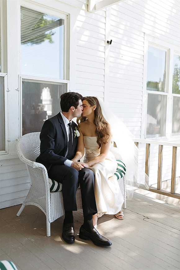 Wedding kiss portrait of bride and groom kissing on a wicker chair, veil draped, holding hands on a white house porch in daylight